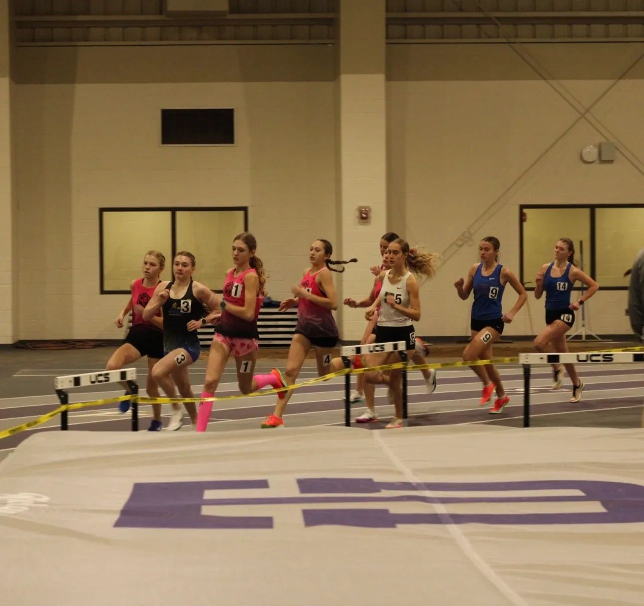 Young female athletes compete in an indoor track race hurdling event.