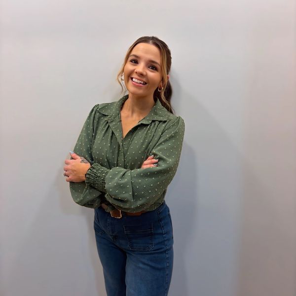 Smiling woman in a green blouse and jeans stands against a plain wall.