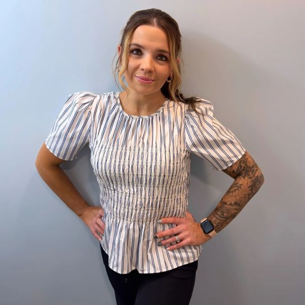 Woman with a striped blouse and tattooed arm poses confidently against a plain wall.