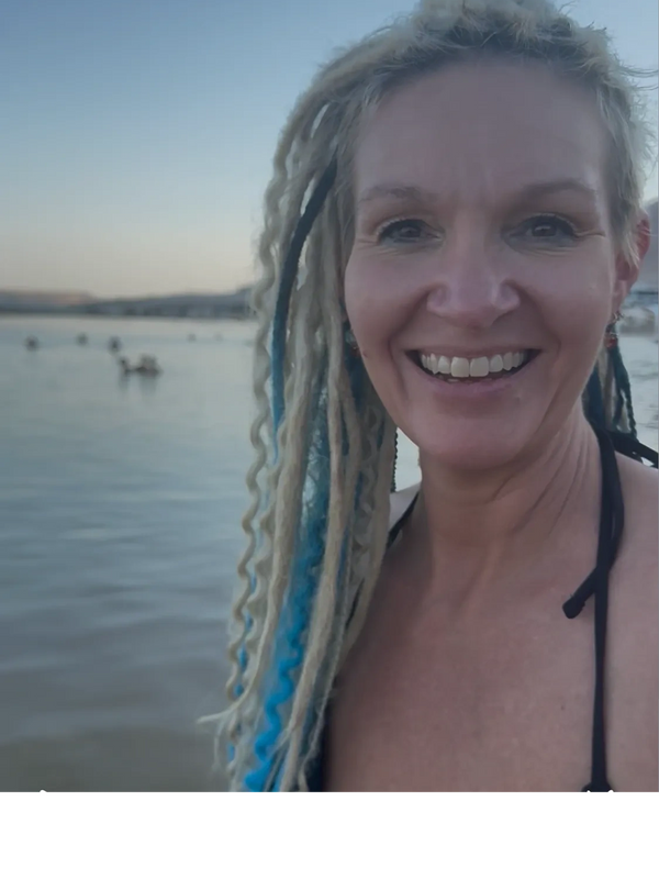 Smiling woman with blue dreadlocks by the water at sunset.