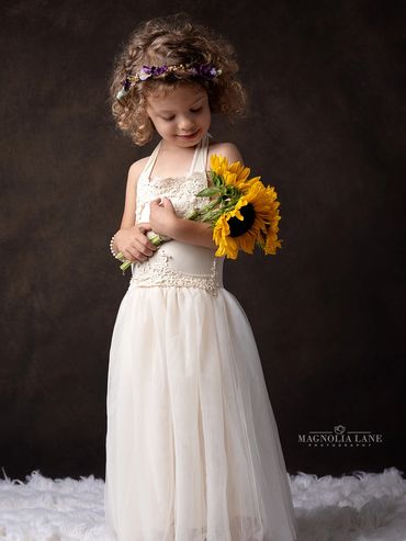 Studio portrait of a toddler girl wearing a long dress on a brown background holding sunflowers.