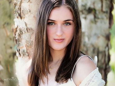 Outdoor portraits young woman with long brown hair wearing white top standing against a tree.