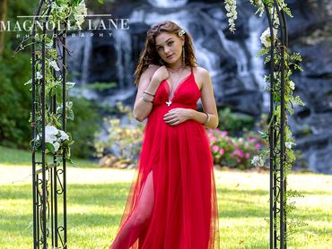 Outdoor maternity portrait of a young woman wearing a red gown with a waterfall behind her.