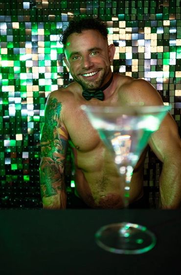 Smiling shirtless bartender with tattoo and bow tie behind a bar with a martini glass.