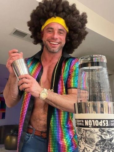 Man in a colorful shirt and afro wig shaking a cocktail shaker with a blender nearby.