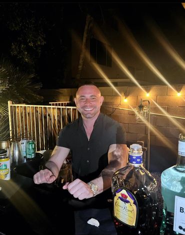 Smiling man behind a bar counter with bottles and string lights at night.