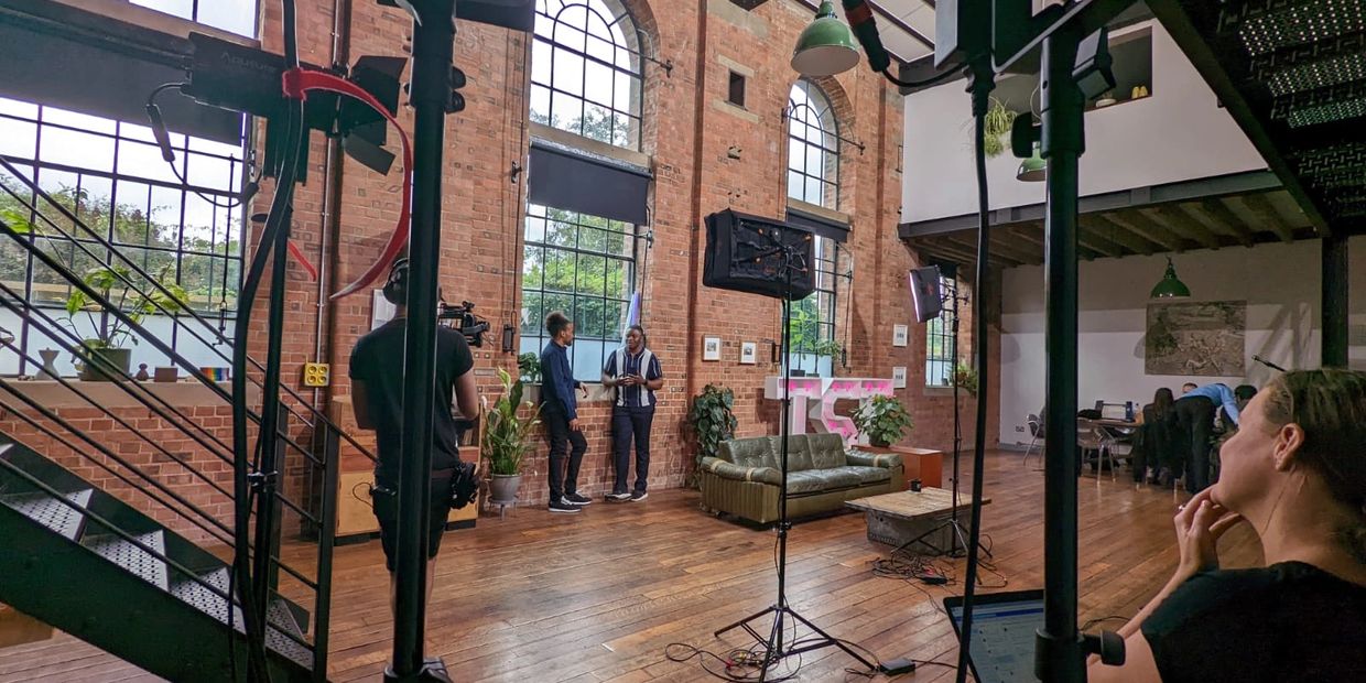 Film crew and hosts in a well-lit studio with brick walls.