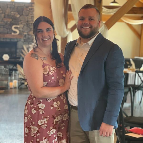 A smiling couple dressed formally at an indoor event with rustic decor.