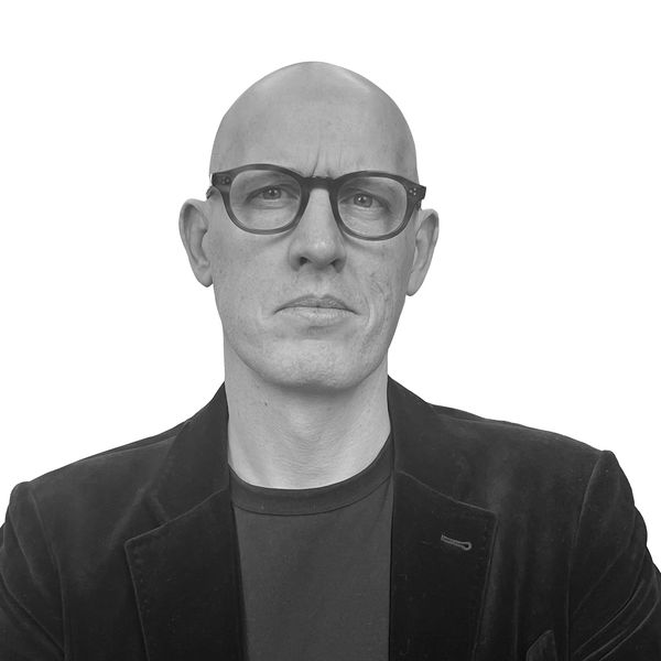 Serious bald man wearing glasses and a black jacket against white background.