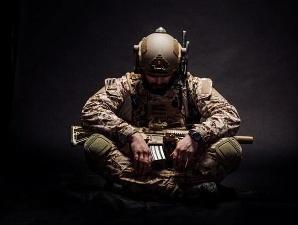 Soldier in tactical gear sits cross-legged holding a rifle in a contemplative pose.