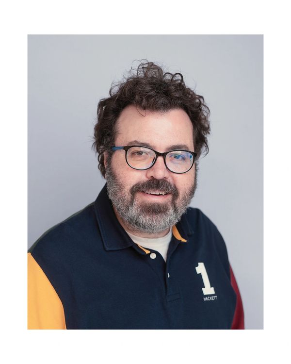 Man with glasses and curly hair smiling against a plain background.