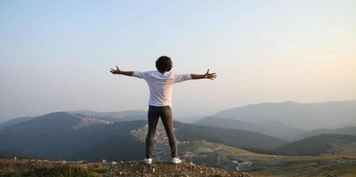 Person standing on a mountain with arms wide open, overlooking a scenic valley.