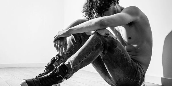 Shirtless man with curly hair sits on floor in jeans and boots, arms resting on knees.