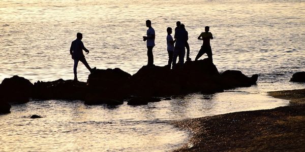 Silhouettes of people standing on rocks by the water at sunset.