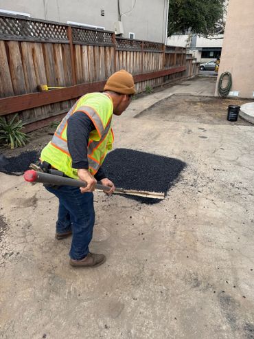 A worker in a safety vest spreads asphalt with a rake on a driveway patch.