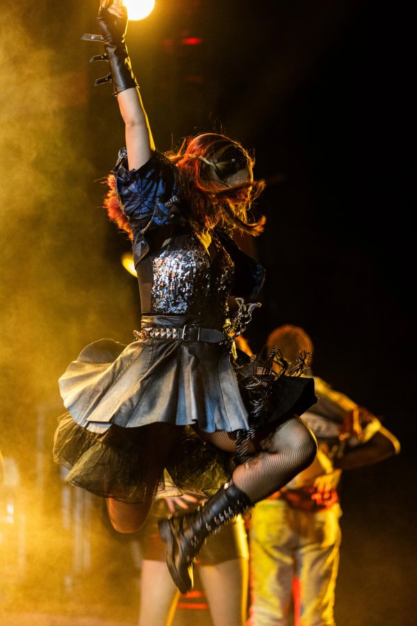 Energetic performer jumps mid-air in dramatic stage lighting and gothic attire.