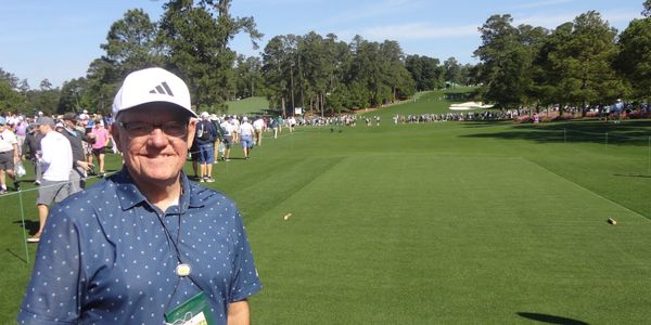 Smiling man at a sunny golf course with spectators walking in the background.