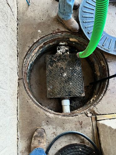 Open manhole with workers' boots and a green hose inside.