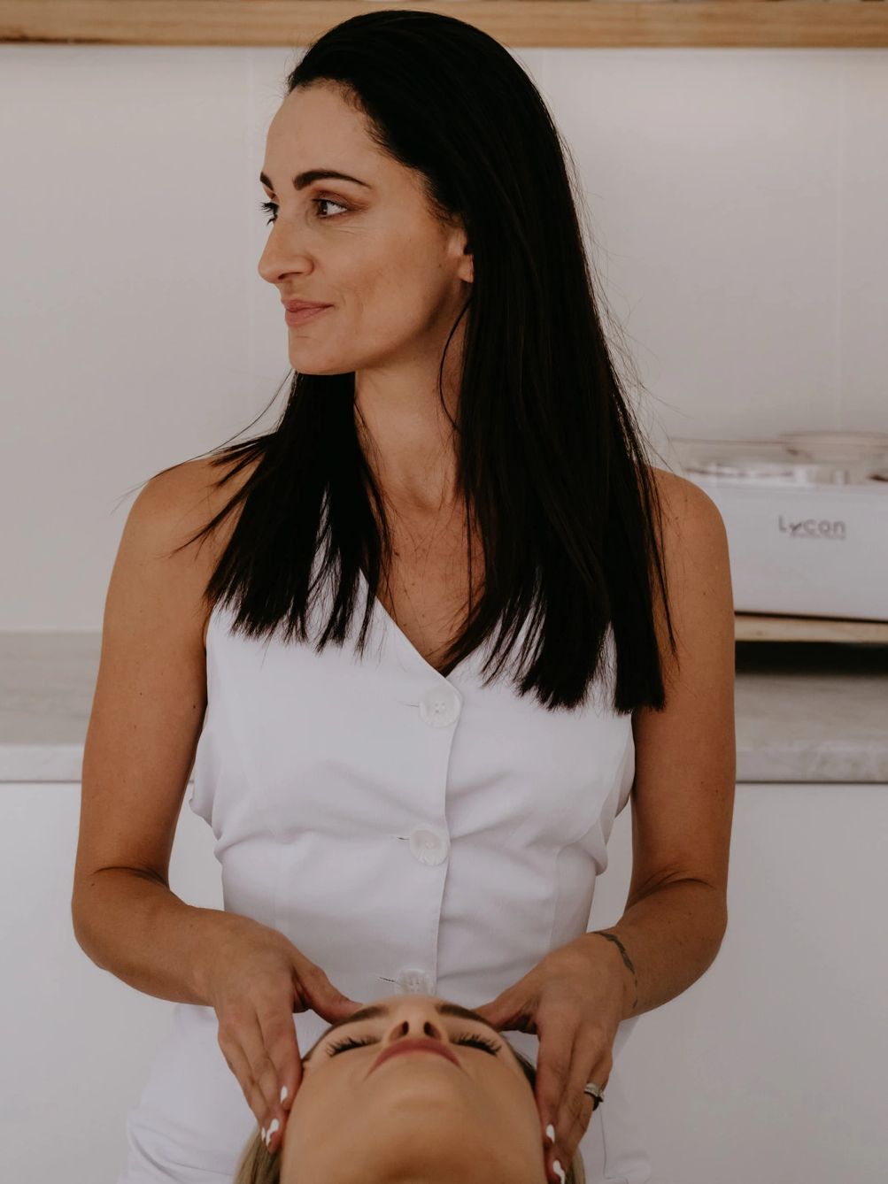 A woman in white performs a gentle facial massage on a lying client in a serene spa setting.