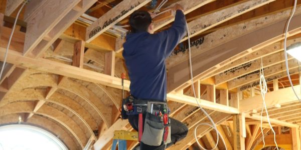 Worker installing wiring on a wooden ceiling structure using a ladder.