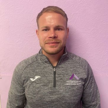 Man wearing a gray sports jacket with a logo, standing against a pink wall.