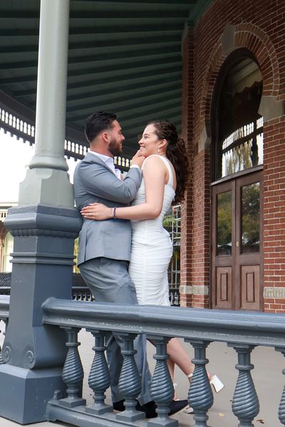 A couple affectionately embracing on a porch with a brick building background.