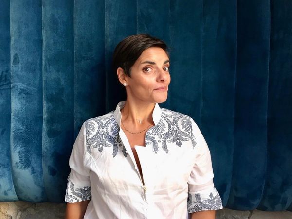 Woman with short hair in a white embroidered shirt against a blue backdrop.