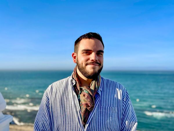 Young man with beard wearing striped shirt and scarf by the ocean under clear blue sky.