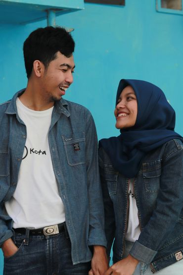 A smiling couple in denim jackets holding hands against a blue wall.