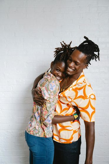 A joyful couple embraces, smiling warmly against a white brick wall.