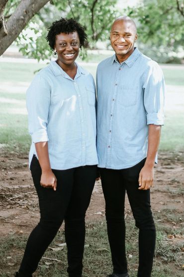 A smiling couple in matching light blue shirts stands under a tree in a park.
