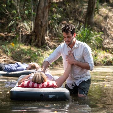A man assisting a woman floating on an air mattress in a shallow river.