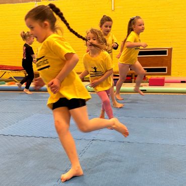 Young girls in yellow shirts running and playing on a blue gym mat indoors.