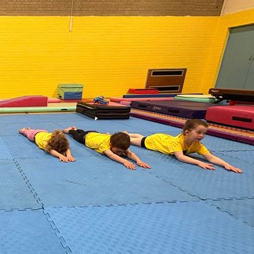 Three children in yellow shirts stretching on blue mats in a gym.