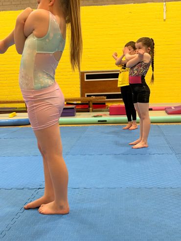 Children practicing gymnastics indoors on blue mats against a yellow wall.