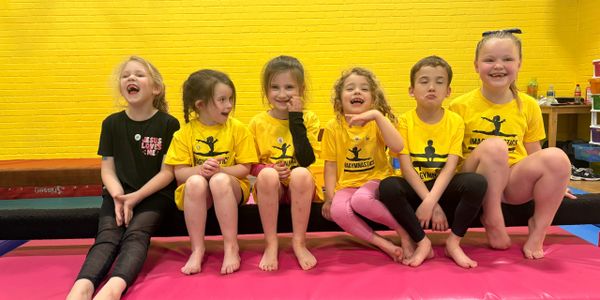 Group of six happy kids sitting on a gym mat against a yellow wall.