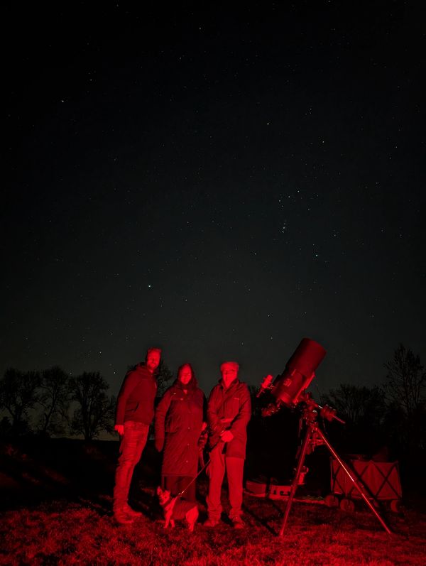 Three people and a dog stargazing with a telescope under a starry night.