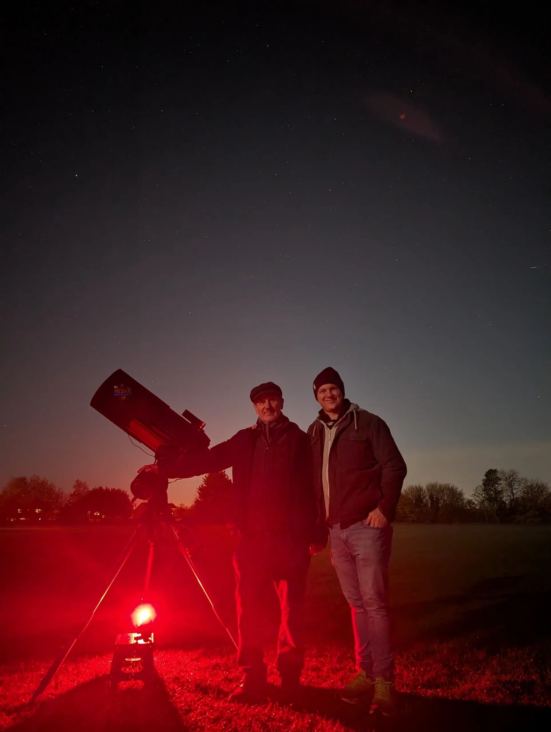 Two men standing by a telescope under a starry night sky illuminated by red light.