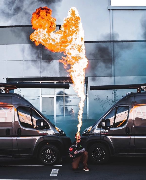 A person performs fire breathing between two black vans in an urban setting.