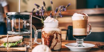 Two fancy coffee drinks and a sandwich on a wooden table in a cozy cafe.