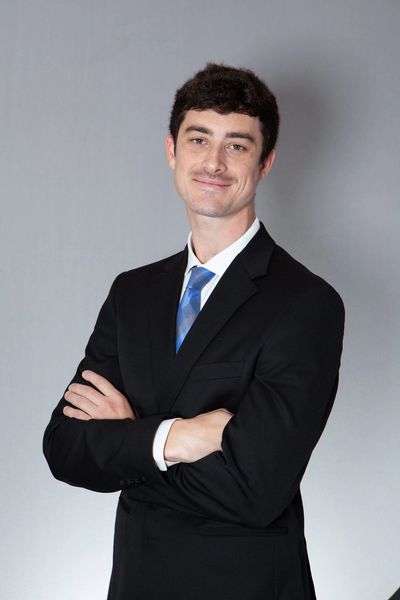 Young man in a black suit with a blue tie, arms crossed, smiling.