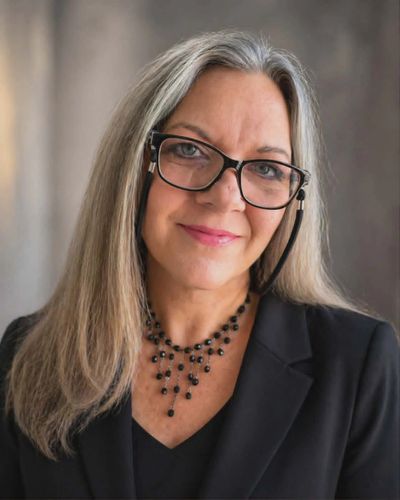 Professional woman with long gray hair and glasses wearing a black blazer.