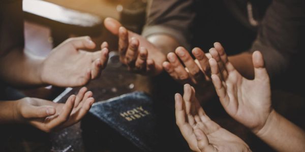Hands raised in prayer around a Bible on a dark surface.