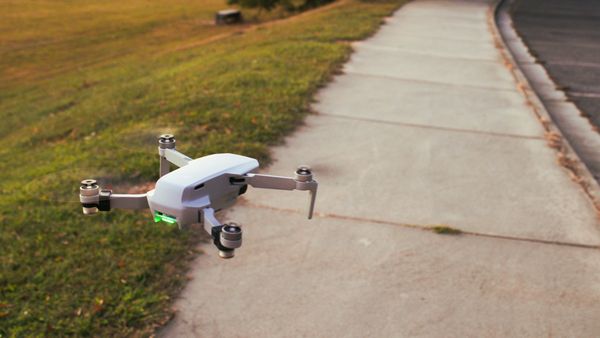 A white drone flying near a sidewalk with a grassy background.
