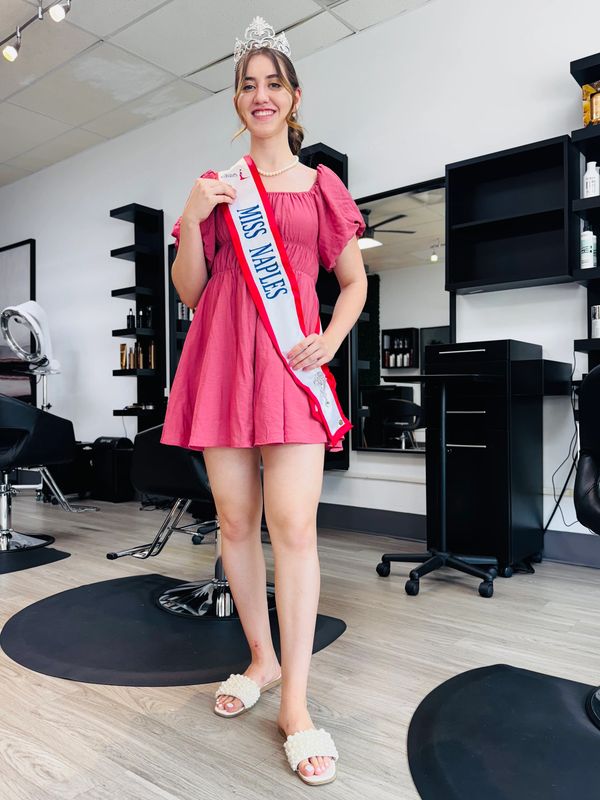 A young woman wearing a Miss Naples sash and crown stands smiling in a salon.