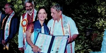 Two individuals holding certificates, wearing ceremonial sashes, smiling outdoors.