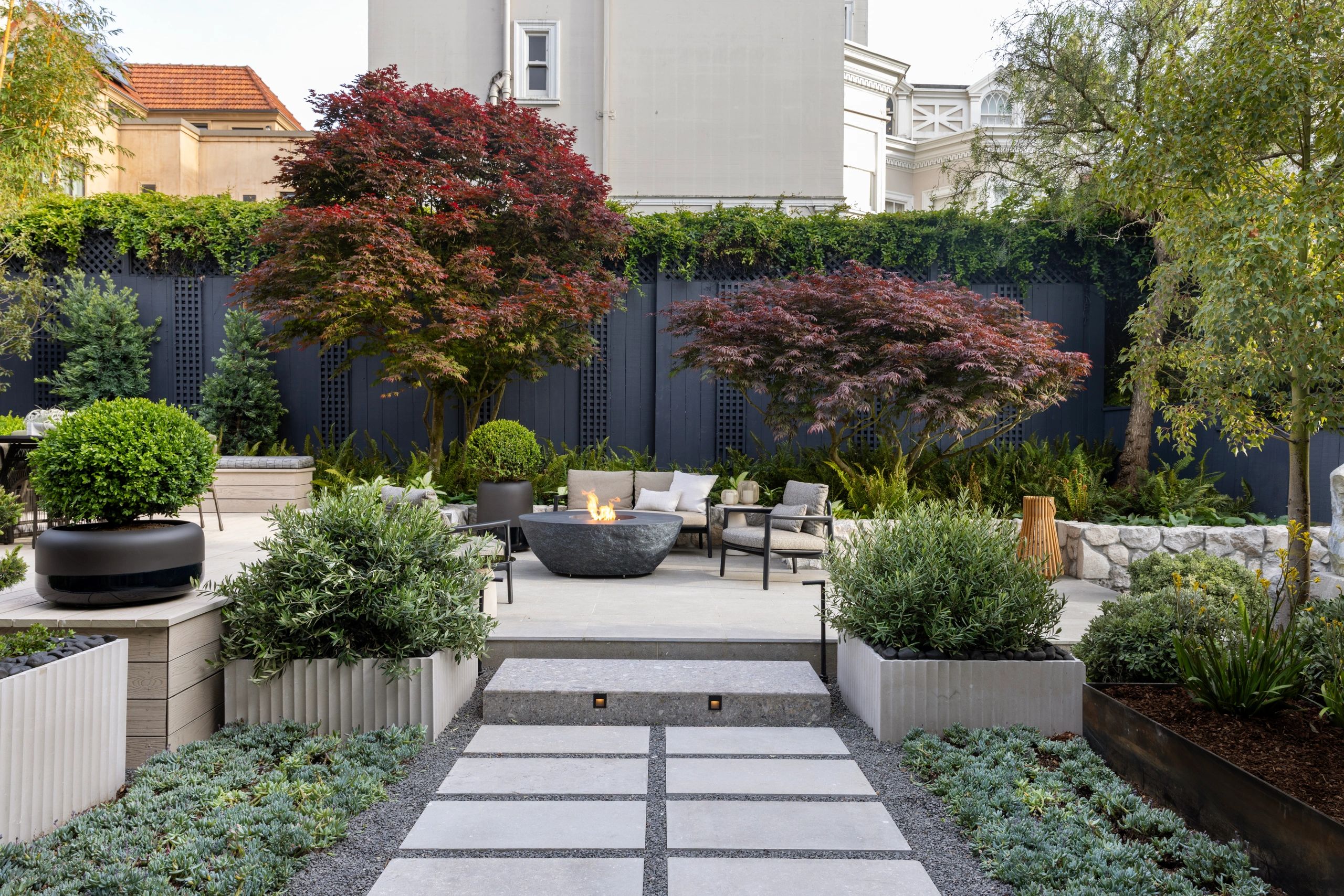 Backyard landscape with red maple trees, light gray hardscaping, furniture, stone planters, and a da