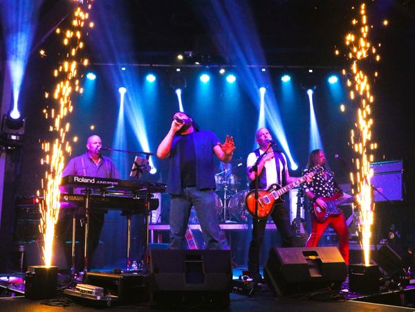 Band performing energetically on stage with bright lights and pyrotechnics.
