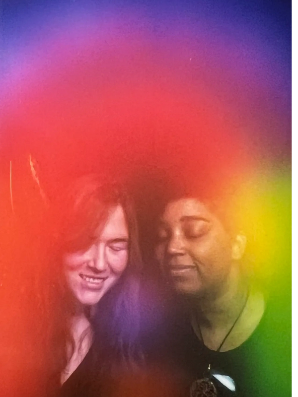 Two women smiling peacefully behind a vibrant rainbow-colored haze.