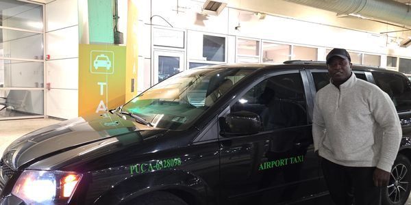 Man standing next to a black airport taxi vehicle inside a parking area.